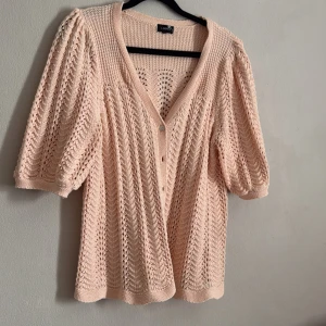 Rosa stickad kofta från Lindex - Säljer en söt rosa stickad kofta från Lindex med v-ringning och knappar framtill. Koftan har trekvartsärmar och ett fint hålmönster som ger en luftig känsla. Perfekt för lager-på-lager-stil!