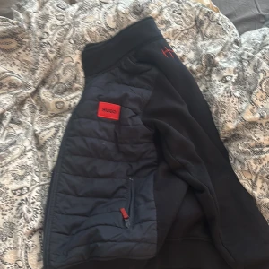 Svart quiltad jacka från Hugo - Säljer en svart quiltad jacka från Hugo med dragkedja och röda detaljer. Jackan har en broderad logga på bröstet och är perfekt för kyligare dagar. Den har långa ärmar och en stilren design.