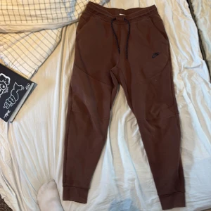 Bruna mjukisbyxor från Nike - Säljer ett par bruna mjukisbyxor från Nike med snörning i midjan och en dragkedja på ena benet. Perfekta för en avslappnad stil. Byxorna har en bekväm passform och är gjorda i mjukt material.