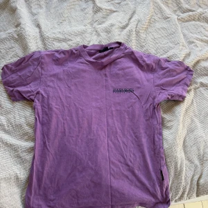 Lila t-shirt från Napapijri - Säljer en lila t-shirt från Napapijri med ett coolt blommönster på baksidan. T-shirten är kortärmad och har en liten logga på framsidan. Perfekt för en avslappnad stil! Den har tre små hål (se sista bilderna) men inget som stör helheten!