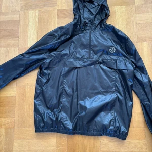 Svart vindjacka från WESC - Säljer en svart vindjacka från WESC med huva och dragkedja. Jackan har en praktisk ficka framtill med dragkedja och elastiska muddar vid ärmslut och nederkant. Perfekt för blåsiga dagar!