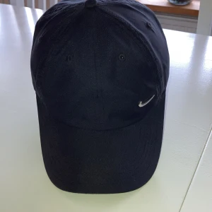Svart keps från Nike - Snygg svart keps från Nike med justerbart spänne baktill. Den har en liten broderad Nike-logga på framsidan. Perfekt för en sportig look eller vardagsstil.
