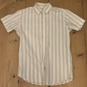 Beige och vit randig skjorta från Jack & Jones - Snygg kortärmad skjorta från Jack & Jones i beige och vit randigt mönster. Skjortan har en klassisk krage och knappar framtill. Perfekt för en stilren och avslappnad look.