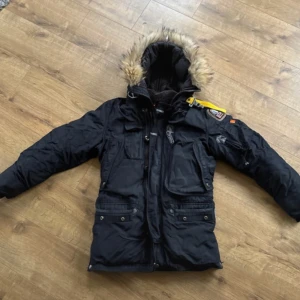 Svart vinterjacka från Parajumpers - Säljer en svart vinterjacka från Parajumpers med pälsfodrad huva och flera praktiska fickor. Jackan har en dragkedja och knappar framtill samt en gul remdetalj på axeln. Perfekt för kalla dagar!