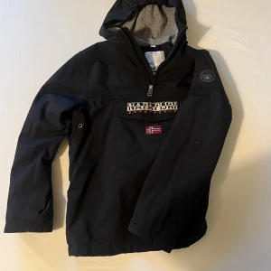 Svart jacka från Napapijri - Säljer en svart jacka från Napapijri med huva och fleecefoder. Jackan har en stor ficka framtill med dragkedja och ett broderat märke med norsk flagga. Perfekt för båda kyliga och varmare dagar! Om du har en fråga så tveka inte på att höra av dig!