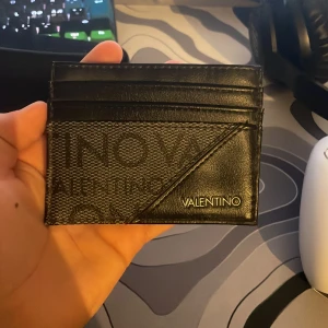 Svart plånbok från Valentino - Snygg och stilren svart plånbok från Valentino med 6st kortfack . Den har en elegant design med en kombination av skinn och ett mönstrat tygparti. Perfekt för att hålla dina kort organiserade och lättillgängliga. Den är som helt ny har nästan inte använt den nån gång 