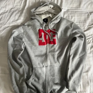 Grå zip up från DC - Säljer en grå hoodie från DC med röd logga på framsidan. Tröjan har dragkedja och justerbar huva med snören. Skriv om det finns några frågor!