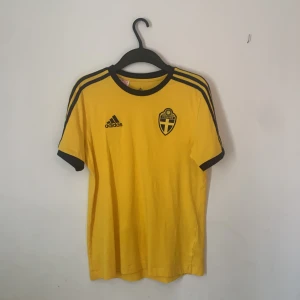 Sverige fotbollströja  - Säljer en gul fotbollströja från Adidas med korta ärmar och svarta detaljer. Tröjan har Sveriges landslagsemblem på bröstet och tre svarta ränder längs axlarna. Perfekt för fotbollsfans!