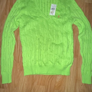 Grön stickad tröja från Ralph Lauren - Säljer en snygg grön stickad tröja från Ralph Lauren med kabelstickat mönster och v-ringning. Tröjan har långa ärmar och en liten logga på bröstet. Fick den som present för ett tag sen men kom aldrig till användning. Perfekt för att lysa upp garderoben! 💚