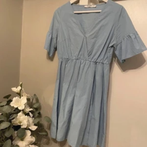 Ljusblå klänning - Säljer en söt ljusblå klänning från Nakd med v-ringning och korta ärmar med volangdetaljer. Klänningen har en avslappnad passform och resår i midjan för en bekväm look. Perfekt för vår och sommar! Storlek S, normal i storlek. Mycket fint skick