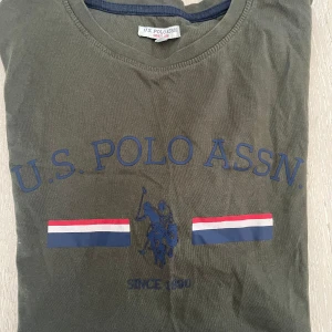 Grön t-shirt från U.S. Polo Assn. - Snygg grön t-shirt från U.S. Polo Assn. med tryck av logotyp och text i blått. T-shirten har korta ärmar och rund halsringning. Rök- och djurfritt hem. Passar 15-16 år (s).