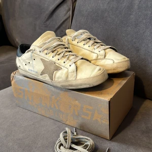 Beige sneakers från Golden Goose - Säljer ett par stilrena beige sneakers från Golden Goose med en ikonisk stjärndetalj i brunt på sidan. Skorna har svarta hälpartier och snörning framtill. Perfekta för en avslappnad look.
