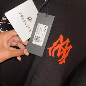 Svart hoodie från Amiri. (Helt ny) - Säljer en svart hoodie från Amiri med en cool orange logga på bröstet och tryck på ryggen. Hoodien har en känguruficka och justerbar huva. Perfekt för en avslappnad stil.