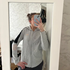 Grå zip up hoodie från Puma - Säljer en grå zip up hoodie från Puma med dragkedja och svarta detaljer på ärmarna. Kan även användas som en träningskofta. Är endast använd några fåtals gånger. 