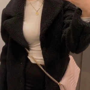 Svart teddyjacka - Mysig svart teddyjacka med långärmad design. Perfekt för kyligare dagar med sin fluffiga och varma känsla. Jackan har en klassisk krage och en stilren look.