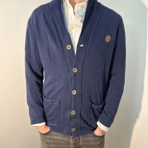 Blå kofta från Marc O’Polo - Säljer en stilren blå kofta med knappar och två fickor framtill. Koftan har en klassisk design med en liten emblem på bröstet och långa ärmar. Perfekt för en avslappnad look. Bra skick, men något urtvättad. Modellen är 182cm/67kg