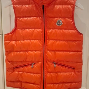 Orange dunväst från Moncler - Snygg orange dunväst från Moncler med dragkedja och två fickor framtill. Perfekt för att hålla värmen med stil. Västen har Monclers ikoniska logga på bröstet. Dragkedjan precis utbytt och det är original färgen på dragkedjan. Original väst i bra skick. Passar för barn mellan som bär storlek 164.