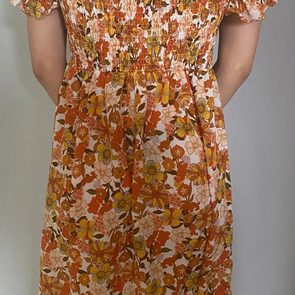 Charmig blommig klänning i varma toner av orange, gul och vit. Klänningen har smockdetaljer upptill och korta puffärmar. Perfekt för en somrig look med sitt lekfulla mönster och bekväma passform.. Mekot.