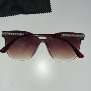 Bruna solglasögon med strassdetaljer - Snygga bruna solglasögon med strassdetaljer på ovansidan av bågarna. De har en gradientlins som går från mörkt till ljust och en elegant design. Perfekta för att ge en glamorös touch till din stil.