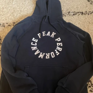 Mörkblå hoodie från Peak Performance - Säljer en snygg mörkblå hoodie från Peak Performance med vit text på framsidan. Den har en klassisk design med huva och dragsko samt långa ärmar. Perfekt för en avslappnad stil.