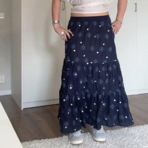 Mörkblå långkjol från Zara - Säljer en mörkblå långkjol från Zara med broderade detaljer och små vita pärlor. Kjolen har en luftig passform och är perfekt för en bohemisk stil. Den är tillverkad i ett lätt och bekvämt material.