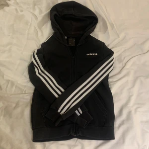 Svart huvtröja från Adidas - Säljer en svart huvtröja från Adidas med klassiska vita ränder längs ärmarna. Tröjan har dragkedja och en bekväm huva. 