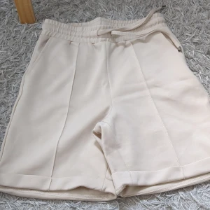 Beige shorts med knytband - Säljer ett par stilrena beige shorts med elastisk midja och knytband. Perfekta för en avslappnad look. De har en enkel design med diskreta sömmar och är gjorda i ett mjukt material.