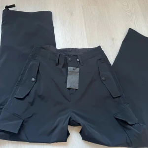 Svarta cargobyxor från J.Lindeberg - Snygga svarta cargobyxor från J.Lindeberg med praktiska fickor och knappdetaljer. Perfekta för en stilren och funktionell look. Tillverkade i ett slitstarkt material. Ordinarie 2000kr