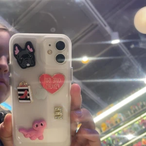 Transparent mobilskal med dekorationer - Säljer ett transparent mobilskal dekorerat med flera roliga detaljer: en svart hund, ett rött hjärta med texten 'Cute but Psycho', en shoppingväska med en röd sko, en glittrig bokstav 'E', och en rosa figur. Perfekt för att ge din mobil en personlig touch!