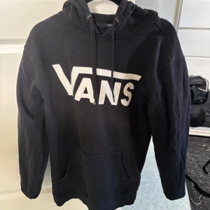 Svart hoodie från Vans - Säljer en svart hoodie från Vans med stor vit logga på framsidan. Tröjan har en klassisk känguruficka och justerbar huva med dragsko. Perfekt för en avslappnad stil.