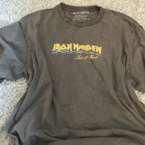 Grå Iron Maiden t-shirt - Säljer en grå Iron Maiden t-shirt med tryck från albumet 'Piece of Mind'. Framsidan har bandets logga i gult och baksidan visar en kedjad figur med låttitlar. Perfekt för fans av klassisk rock! 🤘