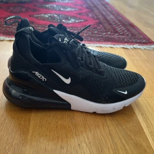 Svarta Nike Air Max 270 - Säljer ett par svarta Nike Air Max 270 sneakers med vit sula och synlig Air-enhet i hälen. Skorna har snörning och Nike-logga i vitt på sidan och tungan. Perfekta för en sportig look!