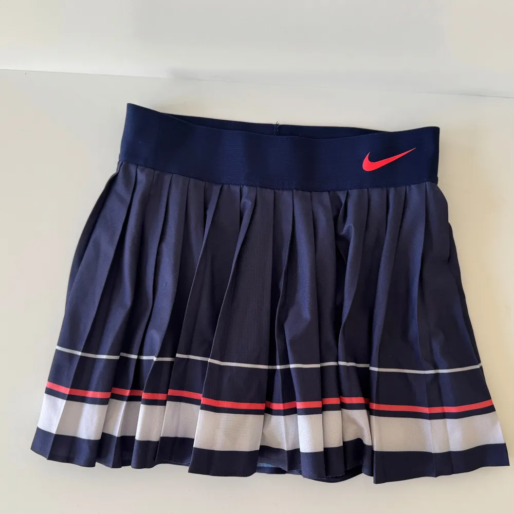 Snygg mörkblå plisserad kjol från Nike med röda och vita ränder. Perfekt för sportiga tillfällen med Dri-Fit material som håller dig sval. Elastisk midja för bekväm passform.. Hameet.
