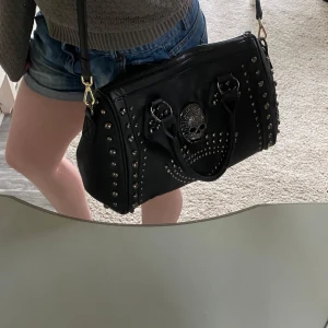 Y2k handväska med döskalle och nitar - Cool svart handväska som jag tidigare köpte på sellpy. Inga defekter. Axelrämsbandet ingår inte eftersom jag har tagit ett från en annan väska och fick inte med ett när jag köpte den tidigare- men det finns hällor. Väskan är lagom stor och har ett dragkedja fack på baksidan, ett likadant fack på insidan samt två tygfack på insidan. Pris kan typ alltid diskuteras! Kolla mina andra annonser också, fråga om du undrar över något!🙌😇