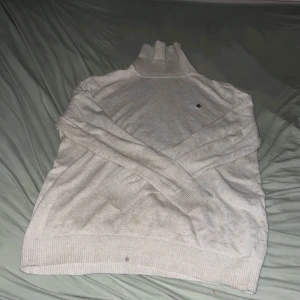 Beige polotröja med broderad detalj - Snygg beige polotröja med ribbade muddar och hög krage. Tröjan har en broderad mörkbrun lilja på bröstet som ger en clean och stilren look. Perfekt för lager-på-lager under kyliga dagar. Materialet är mjukt och stickat för extra komfort.