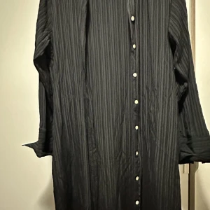 Svart skjortklänning från Lindex M - Svart långärmad skjortklänning från Lindex i storlek M. Klänningen har diskreta vertikala ränder, knappar hela vägen framtill och en avslappnad passform. Perfekt för dig som gillar stilrena och enkla plagg med lite edge.