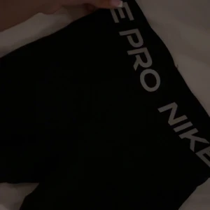 Svarta Nike Pro träningstights shorts - Svarta Nike Pro shorts med tight passform och elastisk midja med logga. Perfekta för träning eller sport, tillverkade i ett stretchigt syntetmaterial som andas. Sitter snyggt och bekvämt på kroppen och ger en sportig look. Dom passar alla möjliga kroppar och längder.