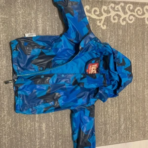 Blå regnjacka Ticket to Heaven 104cm - Blå regnjacka från Ticket to Heaven med coolt geometriskt mönster i olika blå nyanser och svart. Jackan har huva, dragkedja och tryckknappar framtill samt resår vid ärmslut. Perfekt för regniga dagar och lek utomhus.