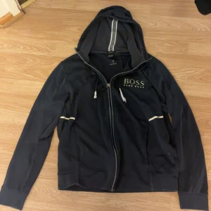 Svart hoodiejacka från Hugo Boss - Svart hoodiejacka från Hugo Boss med dragkedja, vit logga på bröstet och vita detaljer på ärmarna. Jackan har huva med snörning och ribbade muddar. Perfekt för en chill och stilren look.