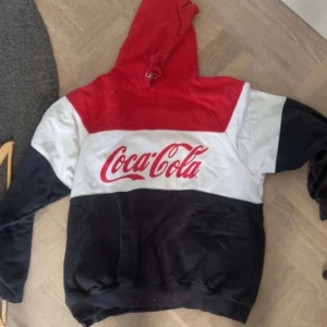 Coca-Cola hoodie i rött, vitt och svart - Säljer en färgstark hoodie från Coca-Cola med stora loggan framtill. Tröjan har huva med snörning och är blockfärgad i rött, vitt och svart. Perfekt för dig som gillar streetwear och vill sticka ut med en ikonisk look.