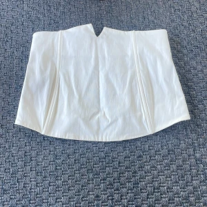 Vit bandeau korsett från NLY Trend - Säljer en vit bandeau korsett från NLY Trend i storlek S. Toppen har en snygg V-formad skärning upptill och smockad baksida för extra komfort. Gjord i ett slätt tyg som känns fräscht och stilrent. Perfekt att styla med jeans eller kjol för en trendig look.