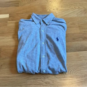 Grå skjorta från Polo Ralph Lauren - Snygg grå skjorta från Polo Ralph Lauren med klassisk krage och knäppning framtill. Skjortan har det ikoniska blå broderade logotypen på bröstet och är tillverkad i mjuk bomull. Perfekt för en clean och stilren look.