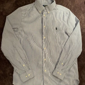 Randig skjorta från Polo Ralph Lauren - Klassisk blå- och vitrandig skjorta från Polo Ralph Lauren med button down-krage och broderad logga på bröstet. Skjortan har långa ärmar, knappar framtill och är tillverkad i bomull. Perfekt för en clean och stilren look. Storlek 16(m)