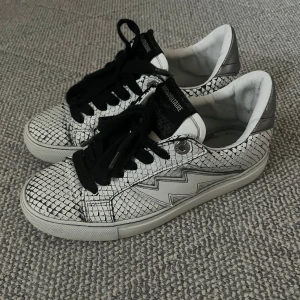 Zadig & Voltaire skor - Snygga sneakers från Zadig & Voltaire i vitt och silver med ormmönstrad yta och svart snörning. Skorna har en cool blixtdetalj på sidan och en platt sula. Perfekta för dig som vill sticka ut med din stil.