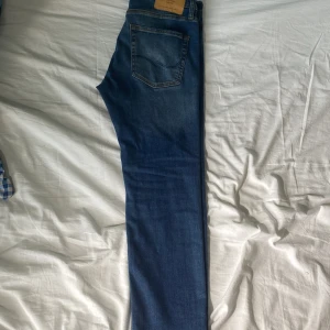 Blå jeans från Jack & Jones - Snygga blå jeans från Jack & Jones med klassisk femficksdesign och knäppning med knappar. Jeansen har en rak passform och är tillverkade i mjukt denimtyg med lite stretch. Perfekta för en avslappnad och stilren look.