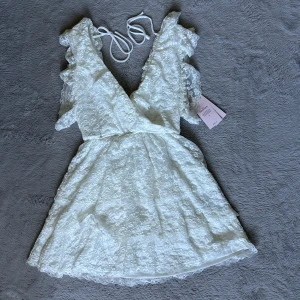 Vit spetsklänning från Nelly  - Superfin vit kort spetsklänning från Nelly med v-ringning, volangdetaljer och omlottkänsla. Klänningen har knytband i ryggen. Aldrig använd utan bara testad, prislapp sitter kvar på klänningen.