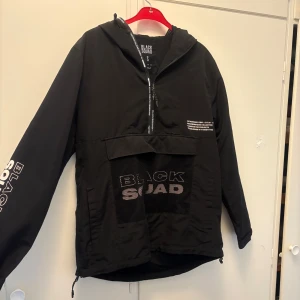Svart vindjacka från Black Squad S - Svart vindjacka från Black Squad i storlek S. Jackan har huva, halv dragkedja framtill och stor meshficka med tryck. Snygga vita logotyper på ärm och bröst. Perfekt streetstil med loose fit och sportig känsla.
