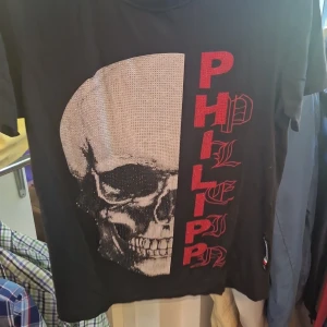 Svart Philipp Plein t-shirt med döskalle STORLEK S - Svart t-shirt från Philipp Plein med stor glittrig döskalle framtill och röd PHILIPP-text i gotisk stil. Klassisk passform med korta ärmar och rund hals. Materialet känns mjukt och skönt, perfekt för dig som gillar edgy streetwear.