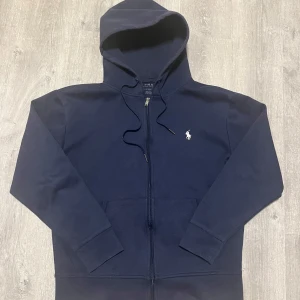 Mörkblå hoodie från Polo Ralph Lauren - Snygg mörkblå hoodie från Polo Ralph Lauren med dragkedja och klassisk broderad logga på bröstet. Tröjan har huva med snörning, känguruficka och är tillverkad i mjuk bomull. Perfekt för en chill och stilren look.