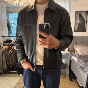 Svart skinnjacka från Tempo - Säljer en klassisk svart skinnjacka från Tempo Leather Collection. Jackan har rak passform, dragkedja framtill, två sidofickor och snygga spänndetaljer i midjan. Perfekt för dig som gillar stilren och tidlös look. Passar till det mesta och är ett måste i garderoben.